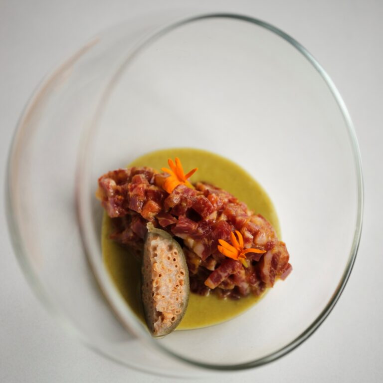 Tartar de Lomo de Presa de Bellota Ibérica FISAN, aliñado con encurtidos, alcaparras, yema asada y un gazpacho piparra con un toque ligeramente picante.